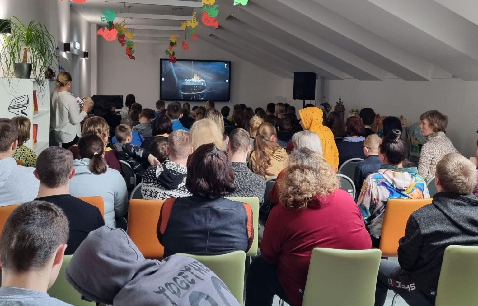 Būrys žmonių iš nugarų stebi filmą ekrane bibliotekos aplinkoje