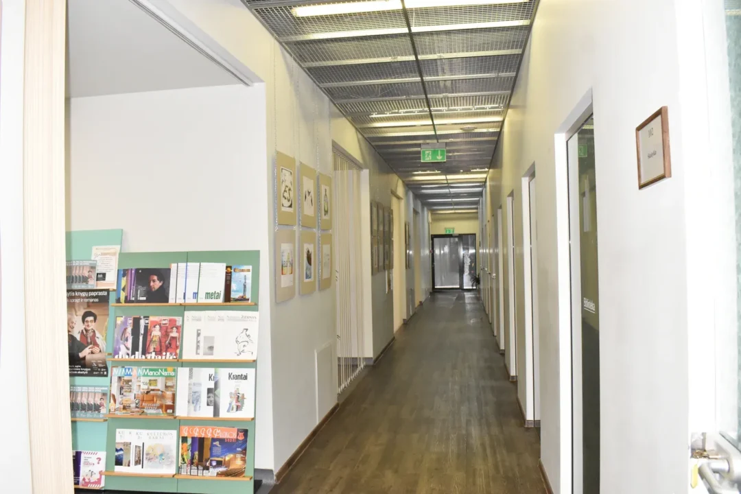 Koridoriuje tamsios rudos grindys, baltos sienos papuoštos paveikslais, daugstiklinių durų.