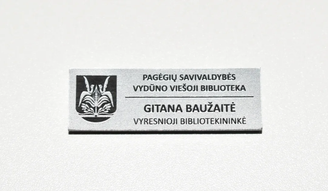 Ženkliukas segamas ant krūtinės, kuriame bibliotekos logotipas ir pavadinimas bei darbuotojo vardas ir pavardė, pareigos.
